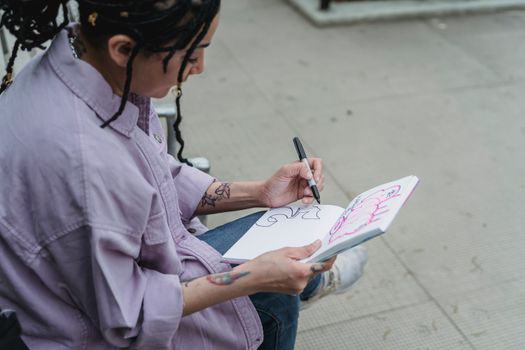 Femme avec des cheveux tressés dessinant dans un carnet de stylisme sur un banc en extérieur.