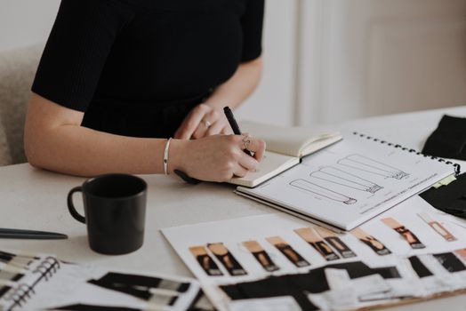 Femme en noir prenant des notes dans un carnet, entourée de croquis de mode et d'une tasse noire.